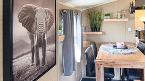Una habitación con una mesa de comedor de madera, sillas, una cortina gris y una estantería con plantas y objetos decorativos.