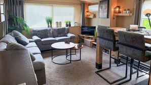 Un salón moderno con un sofá gris, una mesa de centro redonda y un televisor. Hay plantas en macetas y un cuadro en la pared.