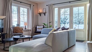 En moderne stue med sofa, lænestol og et lille bord. Store vinduer med gardiner, en plante og en reol med pyngegenstande.