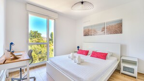 A modern bedroom with a large bed, a desk, and a window with a view of greenery. - Appartement Fleuri, vue mer - Welkeys (Saint-Laurent-du-Var)