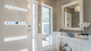 Una habitación con una puerta blanca con paneles de vidrio, un espejo grande con un marco ornamentado, una consola blanca con objetos decorativos, y vistas al exterior a través de la puerta.