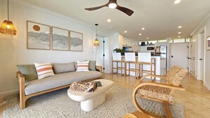 A modern living room with a sofa, coffee table, and woven chairs. There is a kitchen in the background with white cabinets and a bar area.