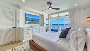 A bedroom with a large bed, a ceiling fan, a TV on the wall, and a balcony with a view of the ocean.