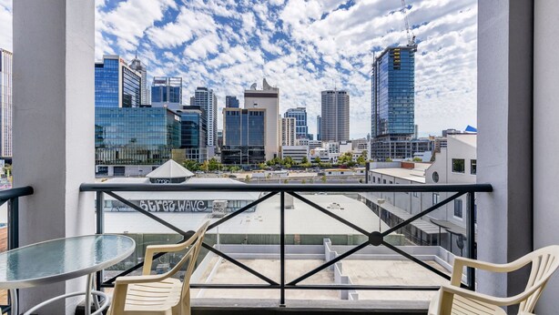A balcony with a glass table and chairs, overlooking a cityscape with modern buildings and a clear sky.