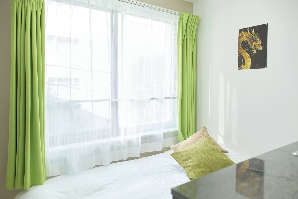 A bedroom with a bed, green curtains, a window, and a wall-mounted artwork.