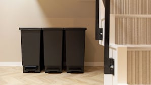 Three black trash bins in a room with a wooden floor and a beige wall.