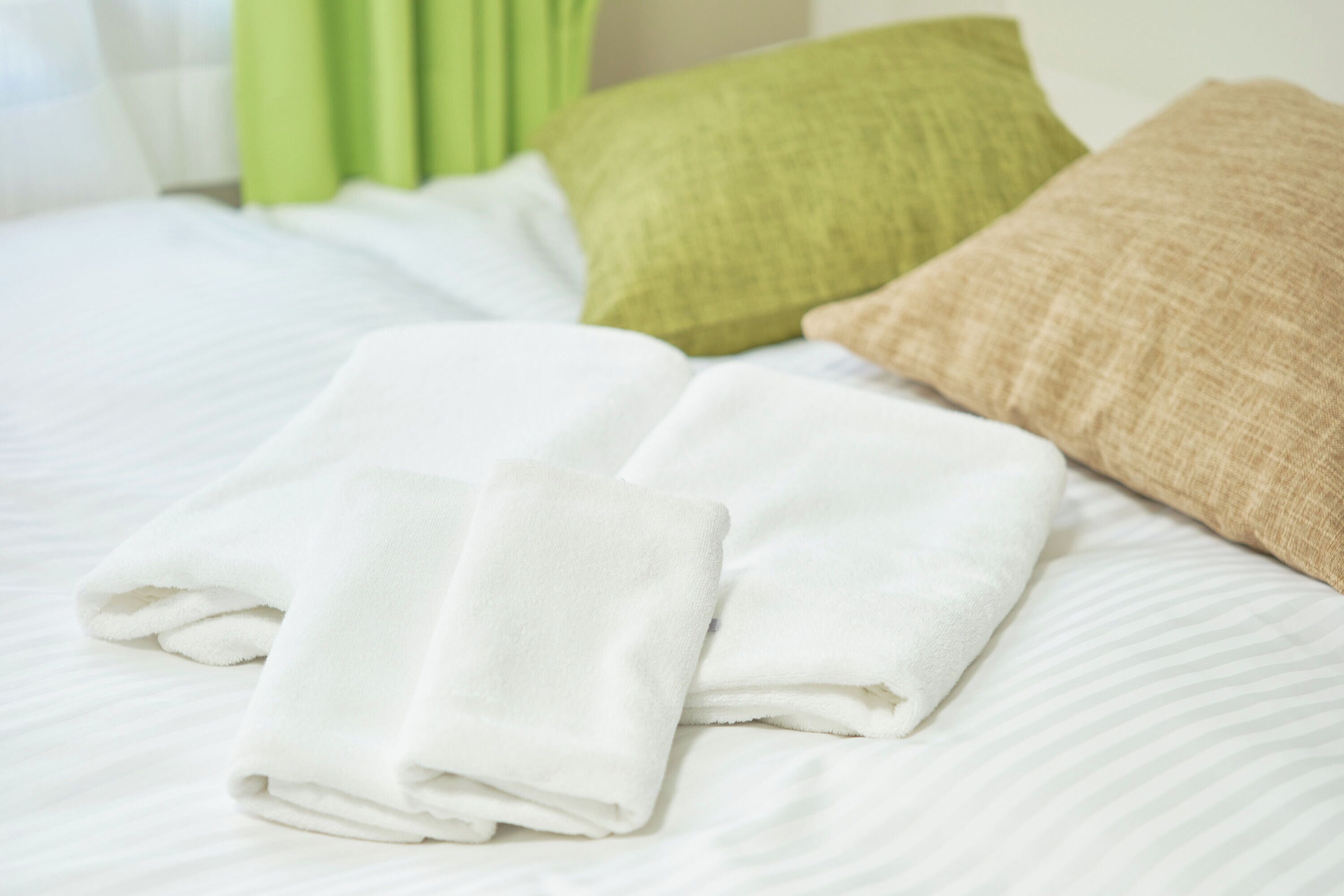 A neatly made bed with white linens, two folded white towels, and two pillows, one green and one beige.