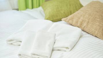 A neatly made bed with white linens, two folded white towels, and two pillows, one green and one beige.