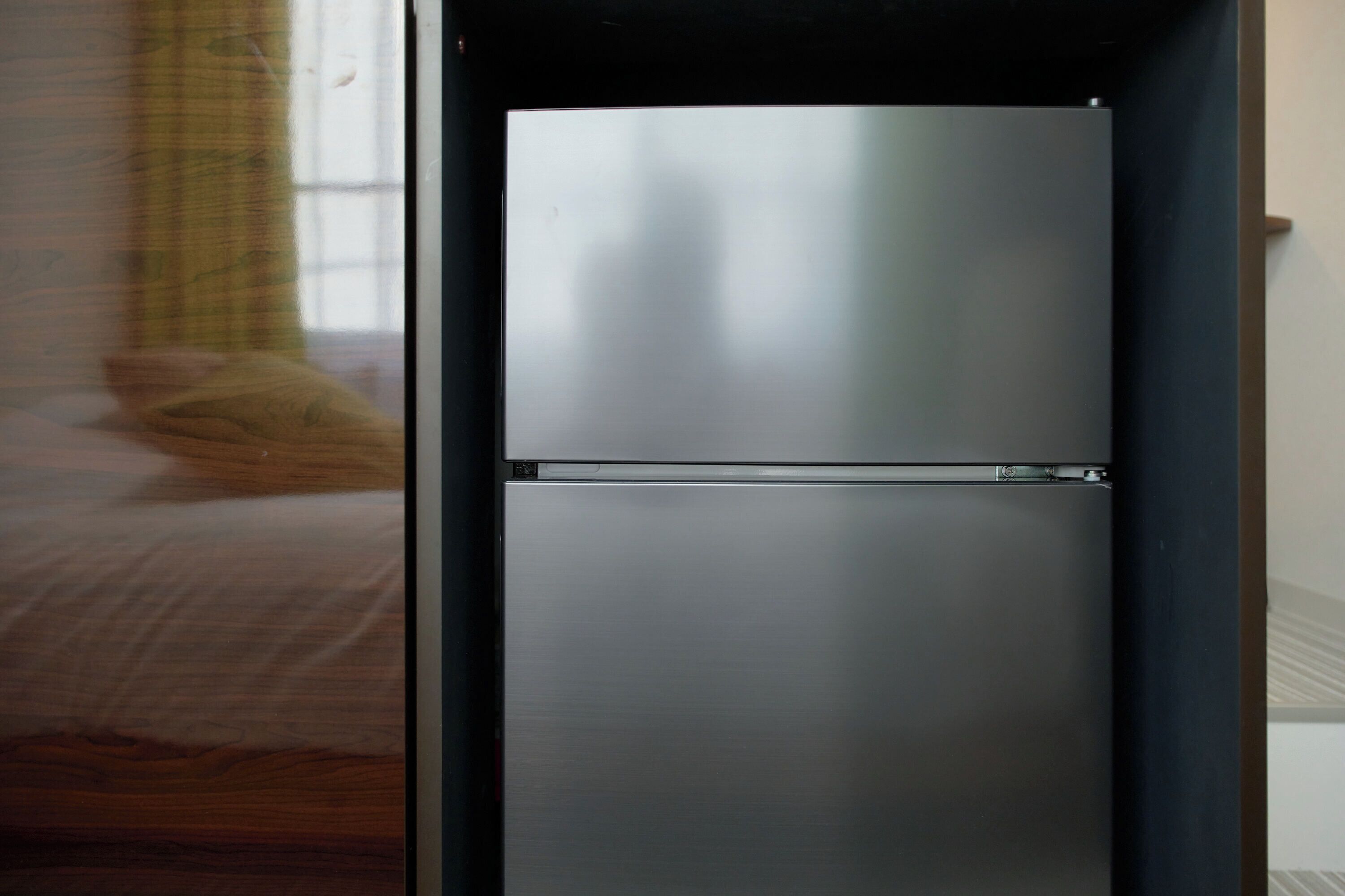 A modern, metallic refrigerator in a room with a wooden floor and a window with a curtain.