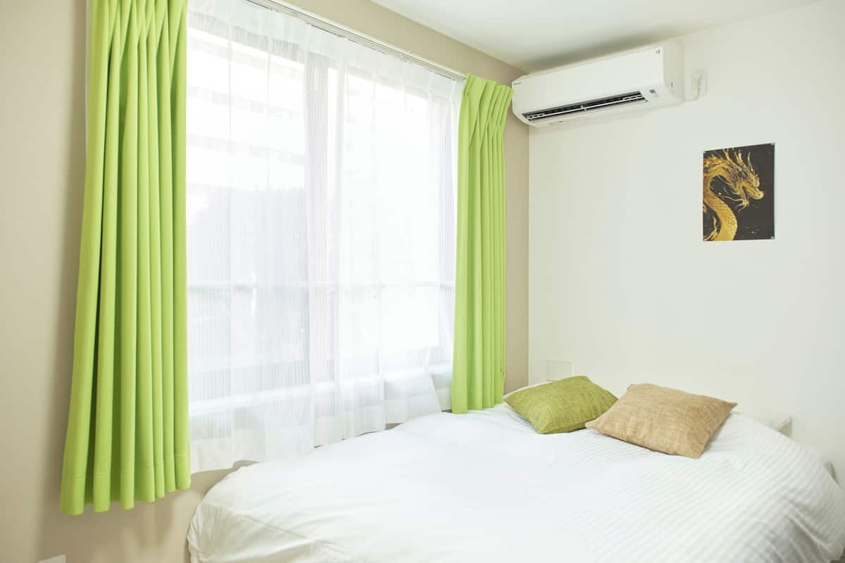 A hotel room with a bed, green curtains, a white air conditioning unit, and a wall-mounted artwork.