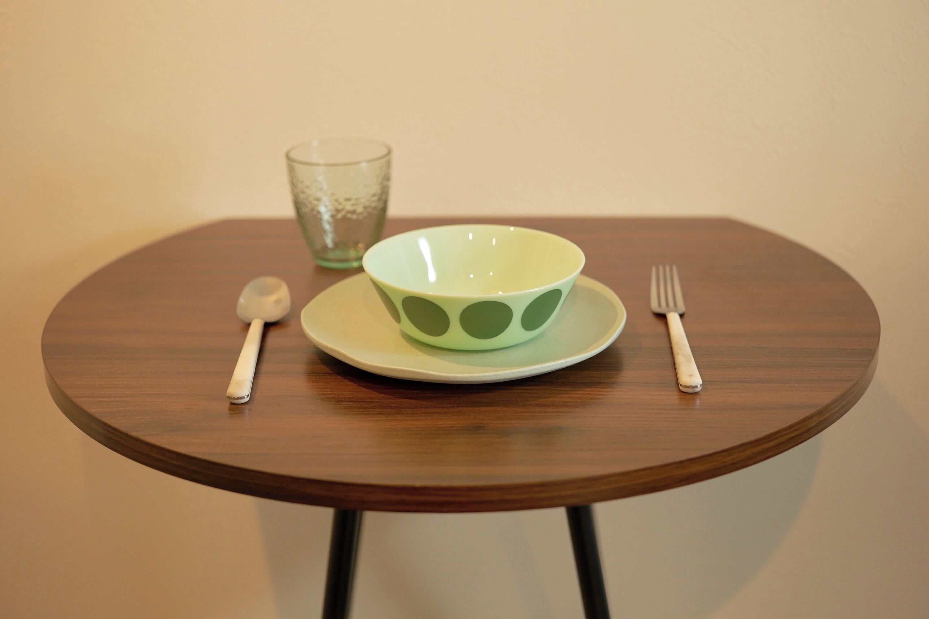 Una mesa de madera redonda con un plato blanco, un cuenco verde con un diseño, una cuchara y un tenedor.