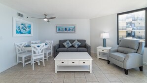 A modern living room with a white coffee table, grey sofa, and white chairs. There are two paintings on the wall, a ceiling fan, and a window with a view of buildings.