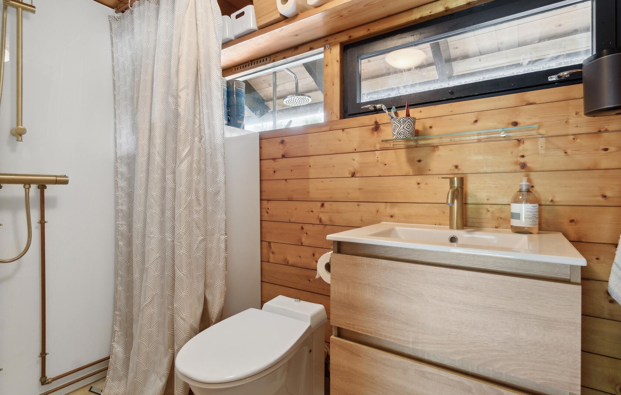 Une salle de bain avec un mur en bois, des toilettes blanches, un lavabo avec un robinet et un espace douche séparé par un rideau.