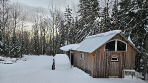 A wooden cabin with a snow-covered roof and a small porch, surrounded by snow-laden trees.