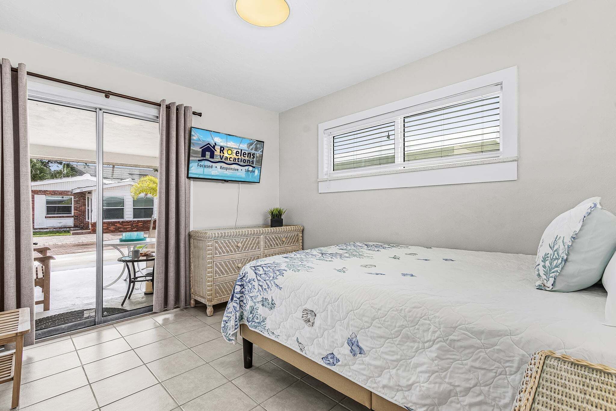 A bedroom with a bed, a television, a window with blinds, and a view of the outside.
