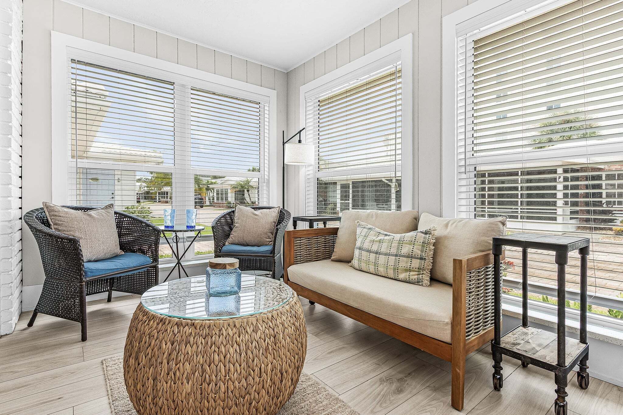 A sunlit room with wicker furniture, a glass coffee table, and large windows with blinds.