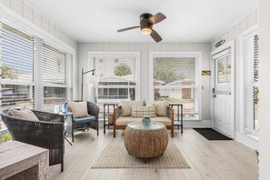 A sunlit patio with wicker furniture, a ceiling fan, and large windows with blinds.