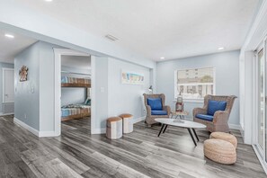 A spacious room with a high ceiling, a bunk bed, a dining area with wicker furniture, and a large window providing natural light.