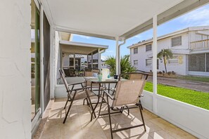 Un patio techado con una mesa y sillas, una puerta de vidrio que da a una cocina y vistas a una calle residencial.