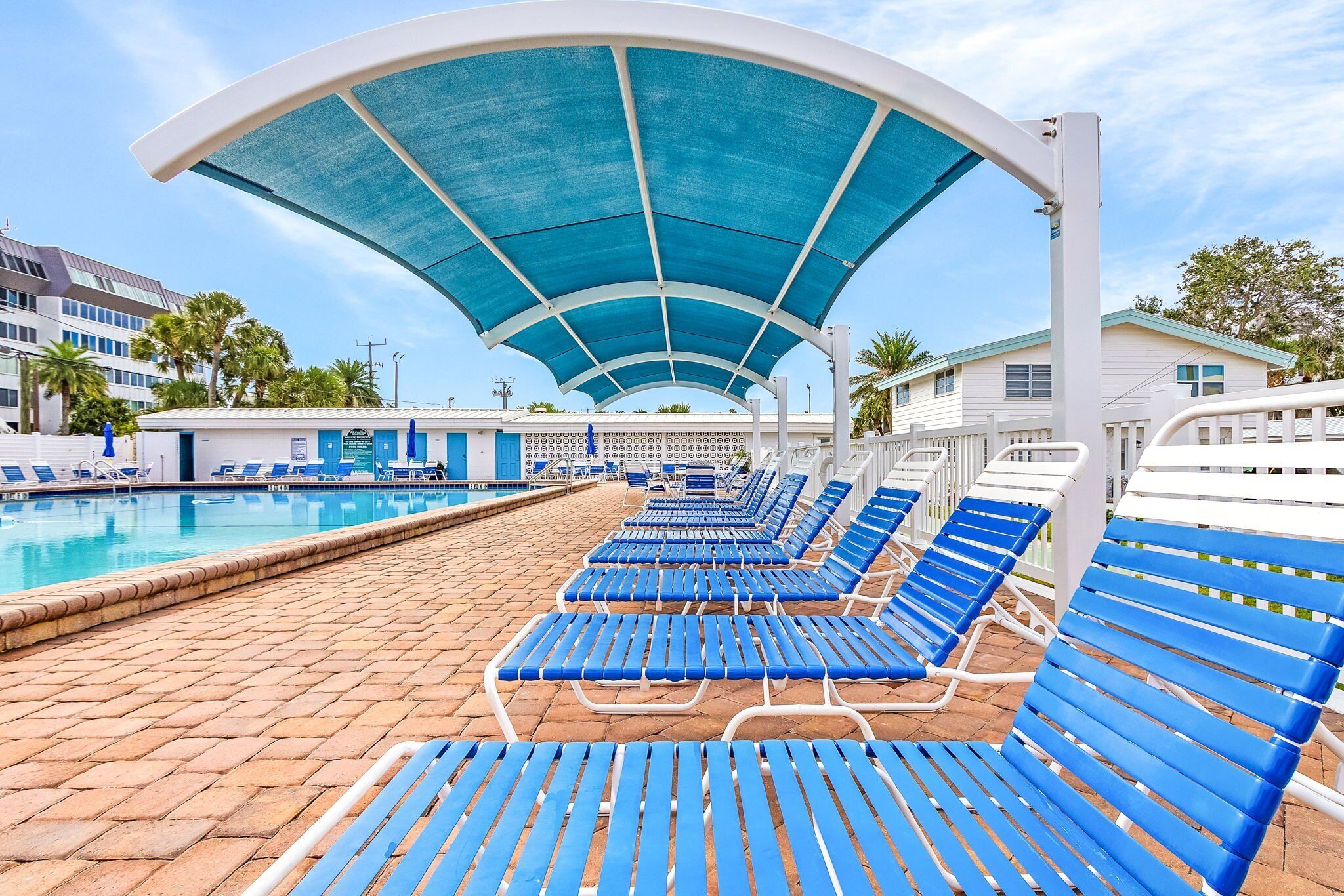 A poolside area with blue lounge chairs under a blue canopy, a pool, and buildings in the background.