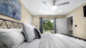 A bedroom with a bed, a ceiling fan, a painting, and a view of the outdoors. - Blumenhaus | Hot Tub | Fire Pit (Fredericksburg)