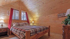 Une chambre à coucher dans une cabane en bois, avec un lit, deux fenêtres avec des rideaux, une table de chevet avec une lampe et une commode.