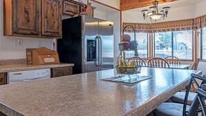 A kitchen with a stainless steel refrigerator, wooden cabinets, granite countertops, and a dining area with a chandelier. - Big Pines Ponderosa Cabin (Overgaard)