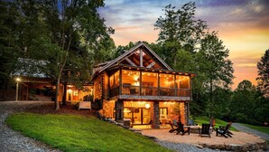 Une cabane en rondins à deux étages, dotée d’un balcon, entourée d’arbres et bordée d’une allée en gravier.