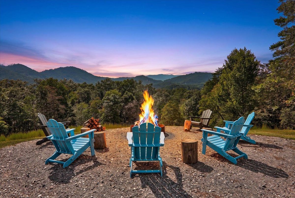 A campsite with three Adirondack chairs arranged around a central fire pit, set against a backdrop of a mountainous landscape.