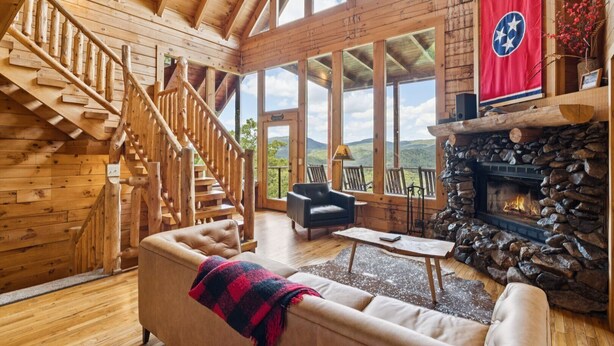 A spacious living room with a wooden staircase, a stone fireplace, and large windows offering a view of the outdoors. - The Cabin - 2 Bedroom with Breathtaking Mountain Views! Hot Tub, and Game Room (Sevierville)