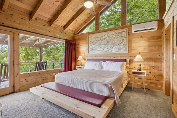 Ein Schlafzimmer einer Holzhütte mit einem großen Bett, zwei Nachttischen, einer Karte an der Wand und einem Balkon mit Blick auf Bäume.