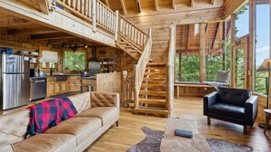 A spacious wooden cabin with a kitchen, a living area with a sofa and armchair, a staircase, and large windows offering views of the outdoors. - The Cabin - 2 Bedroom with Breathtaking Mountain Views! Hot Tub, and Game Room (Sevierville)