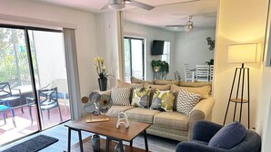 Un salon moderne avec un canapé, une table basse et des fauteuils. On aperçoit un balcon avec du mobilier d’extérieur à travers la porte vitrée.