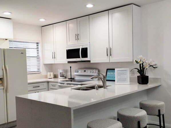 A modern kitchen with white cabinetry, a built-in microwave, a stove, and a sink. There are three stools at the island and a potted plant on the counter.