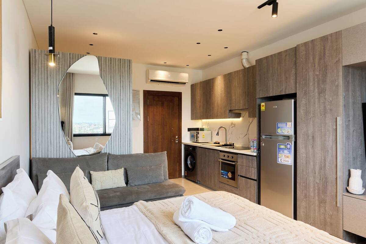 A modern hotel room with a bed, a sofa, a kitchen area, and a large mirror.
