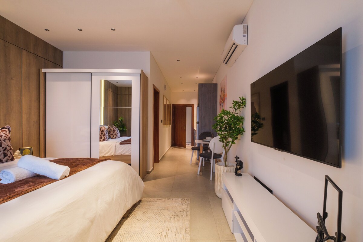 A modern hotel room with a large bed, a flat-screen TV mounted on the wall, a desk with a chair, and a small potted plant.