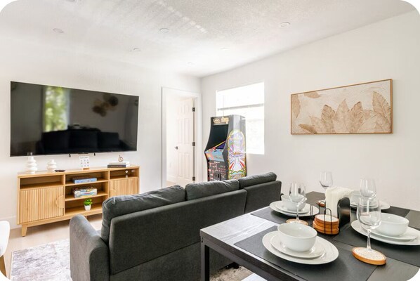 A modern living room with a large flat-screen TV, a gray sofa, a wooden entertainment unit, and a dining area with white plates and glasses.