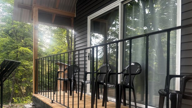 Una terraza de madera con barandilla de metal, sillas de plástico negro y un ventanal con vista a un área boscosa.