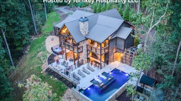 A large, multi-story lodge with a hot tub and outdoor seating area, surrounded by dense forest.