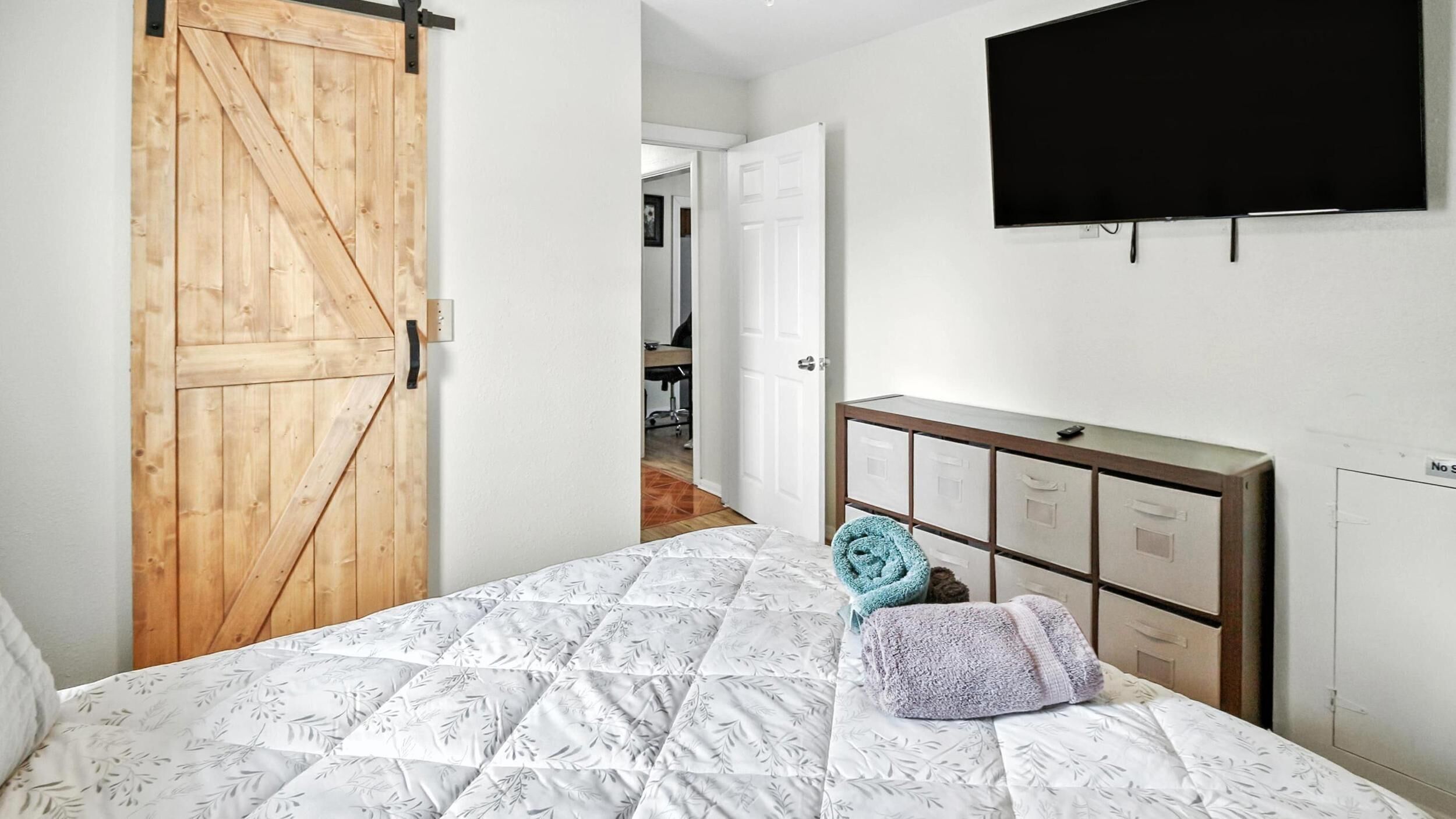 Une chambre avec une porte en bois, un lit avec une literie à motifs, une commode et un téléviseur fixé au mur.