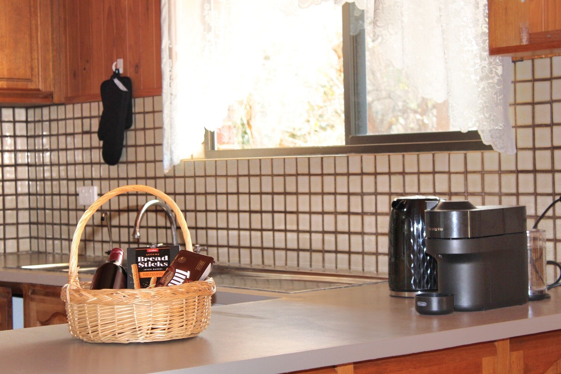 A kitchen with a woven basket containing bread sticks and a bottle, a coffee maker, and a kettle on the counter.