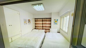 A small, clean room with two single beds, a wardrobe, a window with curtains, and a ceiling light.