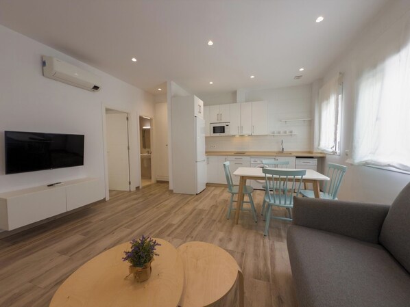 Un apartamento moderno con televisor de pantalla plana, zona de comedor con sillas, sofá, cocina con armarios blancos y piso de madera.