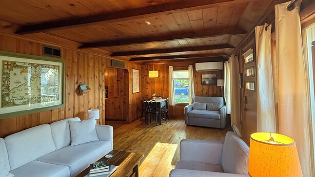 Interior de una cabaña con paredes y techo de madera, un sofá, una mesa y una lámpara.