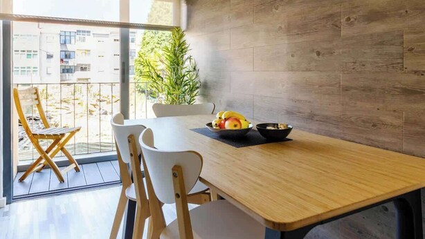 Comedor con mesa de madera, sillas blancas y vistas a edificios a través de grandes ventanales.