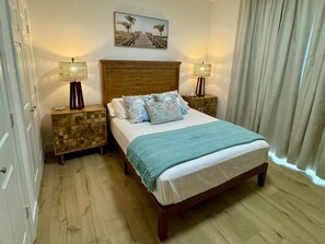 Une chambre à coucher avec un lit en bois, deux tables de chevet, une commode en bois, un tableau sur le thème de la plage et une fenêtre avec des rideaux.
