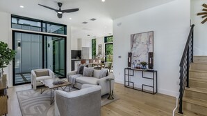 A spacious living area with a modern design, featuring a sofa set, a coffee table, and a dining table. There is a staircase leading to an upper level, and a large window providing natural light. - Stunning Modern Zilker Home with Private Pool, Designer Touches & Prime Location (Austin)