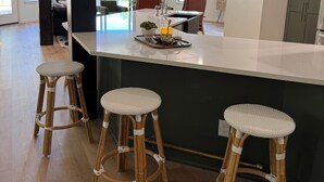 Una cocina moderna con una isla central, tres taburetes de mimbre, iluminación colgante y vistas al exterior a través de amplios ventanales.