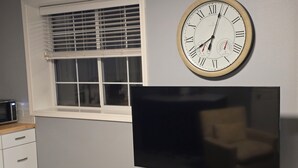Ein Zimmer mit Flachbildfernseher auf einem Holzboden, einer Wanduhr, einem Fenster mit Jalousien und einem weißen Schrank mit Schubladen.