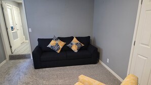Ein Wohnzimmer mit einem schwarzen Sofa, zwei Kissen mit Muster und einem gelben Hocker.